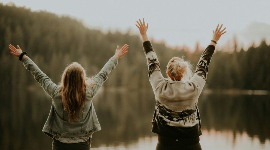 two women hands up standing beside body of water