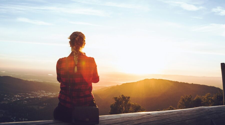 person sitting on wood fence facing mountain under cloudy sky